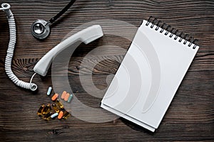 Phone handset, pills, phonendoscope and notebook on dark wooden desk top view call doctor mock up