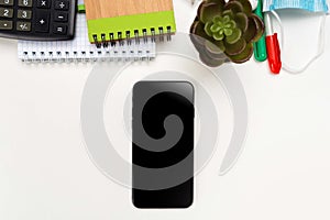 A phone with a screen mockup, a coffee cup of writing supplies, a pens, a notepad on a white wooden table background. Top view of