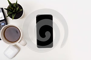 A phone with a screen mockup, a coffee cup of writing supplies, a pens, a notepad on a white wooden table background. Top view of