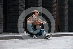 With phone. Stylish man with beard in khaki colored jacket and in jeans is outdoors near building