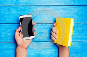 Phone versus book. A man is holding a yellow book and a phone on a blue wooden background. The choice between study and phone. Tel