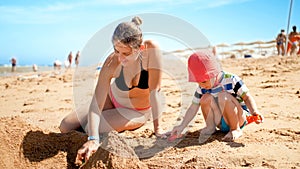 Photo of 3 years old toddler boy building sand castle with young mother the ocean beach. Family relaxing and having fun