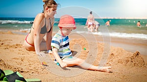 Photo of 3 years old toddler boy building sand castle with young mother the ocean beach. Family relaxing and having fun