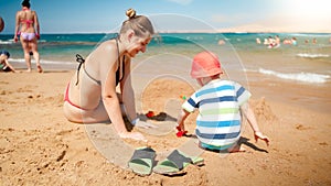 Photo of 3 years old toddler boy building sand castle with young mother the ocean beach. Family relaxing and having fun