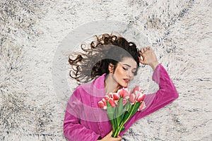 Photo from above of beautiful woman wearing girlish housecoat holding flowers while lying on white fur in apartment photo