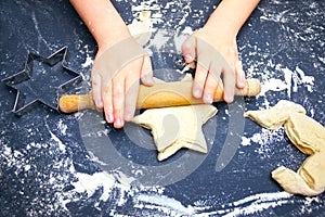 A photo from above of Kid`s hands, some flour, wheat dough and rolling-pin on the black table. Children hands kneading the star d