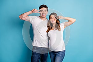 Photo of adorable cheerful attractive couple expecting child showing v-sign cover eye isolated blue color background
