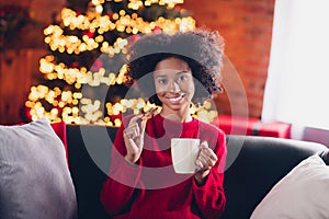 Photo of adorable cheerful little girl sit couch enjoy baked cookie tea mug x-mas decoration lights indoors