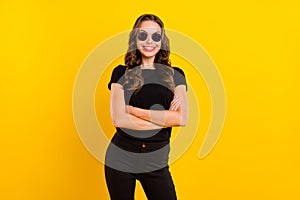 Photo of adorable confident young lady dressed black clothes dark eyewear arms crossed smiling isolated yellow color