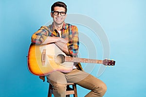 Photo of adorable cute guy dressed plaid shirt spectacles smiling siting chair holding guitar empty space isolated blue