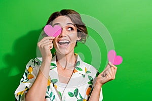 Photo of adorable excited girl dressed print shirt holding two pink hearts cover eye looking empty space isolated green