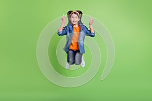 Photo of adorable funky preteen girl dressed jeans shirt showing v-sign jumping high smiling isolated green color