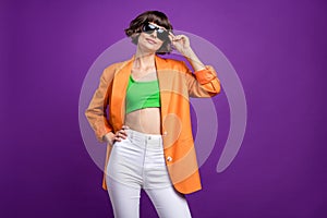 Photo of adorable shiny young woman dressed orange blazer arm dark glasses empty space smiling isolated purple color