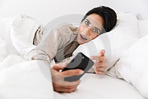 Photo of adorable woman 30s using smartphone, while lying in bed with white linen at home