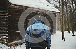 Photo of adventurer hiker in snow covered forest in front of abandoned cottage view from his back. Hiker Man in blue jacket