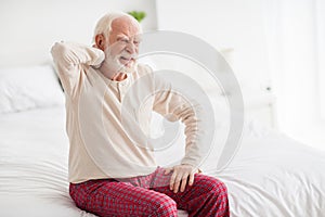 Photo of aged man pensioner sit on bed house indoors unhappy sad upset hand touch shoulders pain spasm ache