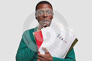 Photo of aghast stupefied dark skinned guy keeps hand on chest, analyzes information from paper documents, studies