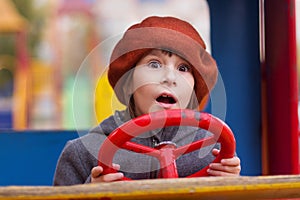 Photo of amazed girl in cap keeping red steering wheel on a playground. Close up portrait of little girl opening mouth and
