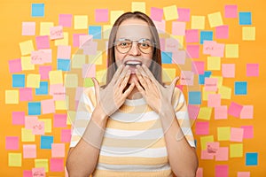 Photo of amazed surprised woman with brown hair in striped t shirt standing against yellow wall with colorful stickers, sees