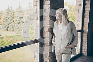 Photo of amazing blond adorable aged granny homey good mood looking dreamy to window standing near brick wall wear