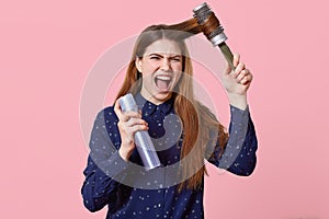 Photo of annoyed young woman combs her hair, dissatisfied with shampoo, holds hairbrush and hairspray, looks with irritated