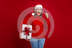 Photo of astonished cheerful granny hold giftbox rejoice victory wear hat pullover isolated red color background