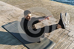 Photo of athletic sportsman 20s in tracksuit lying on fitness mat and doing stretching exercise by seaside