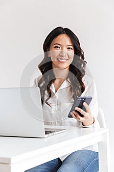 Photo of attractive asian office woman with long dark hair sitting at table and holding smartphone while working with laptop, iso
