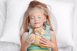 Photo of attractive female child has pleasant dreams at night, holds teddy bear, dressed in pyjamass, sleeps in bedroom on comfort