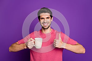 Photo of attractive handsome guy good mood raise thumb up toothy smiling hold hot coffee cup wear casual pink t-shirt