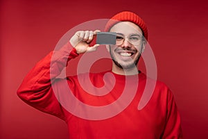 Photo of attractive man with beard in glasses and red clothing. Happy man holds credit card in front of face, 