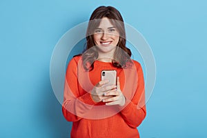 Photo of attractive woman, looks smiling at camera and holding mobile phone, isolated over blue background, winsome lady using