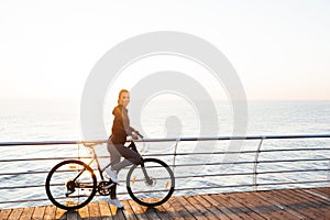 Photo of attractive woman riding bicycle on boardwalk, during sunrise over ocean