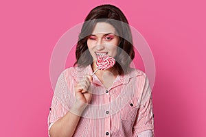 Photo of attractive young woman with dark haired woman with bright makeup, bites candy, blinks eye, poses over pink background,