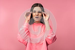 Photo of attractive young woman in glasses looks unexpectedly and surprised, wears casual pink t-shirt white pants