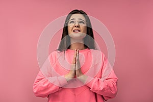 Photo of attractive young woman in glasses uncertainly praying , wears casual pink t-shirt isolated pink color