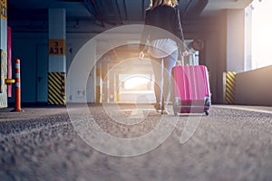 Photo from back of blonde with pink suitcase walking along passage at airport, blurred background