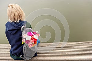 Photo from the back of young blonde dreamy woman with bouquet of flowers in grey backpack near a river. romantic mood