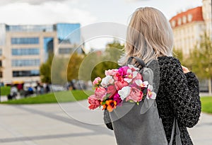 Photo from the back of young blonde romantic woman with bouquet of flowers in grey backpack, standing on a street