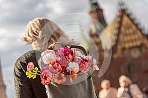 Photo from the back of young blonde romantic woman with bouquet of flowers in grey backpack, standing on a street