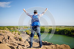 Photo from back of young tourist with backpack with hands up against background of mountain landscape, lake
