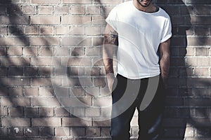 Photo bearded man with tattoo wearing blank white tshirt and black jeans. Bricks wall background. Wide mockup