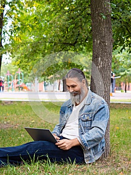 Photo of a bearded man while working online with a laptop. He sits in the park on the grass and works remotely.