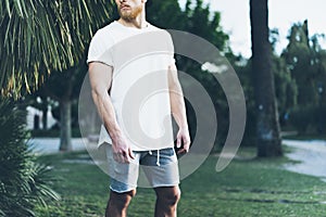 Photo Bearded Muscular Man Wearing White Blank t-shirt and shorts in summer time. Green Tree Palm, Blurred Background