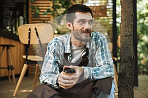 Photo of bearded waiter man sitting on wooden floor while working in cafe or coffeehouse outdoor