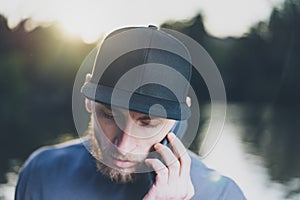 Photo Bearded Young Man Wearing Black Blank Cap Using Modern Smartphone. Green City Park Lake Background and Sunset