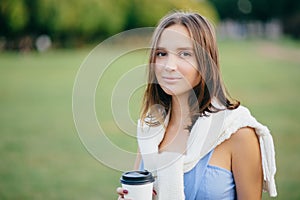 Photo of beautiful comely girlfriend with white sweater on shoulders, drinks takeaway coffee, thinks about something, stands outdo