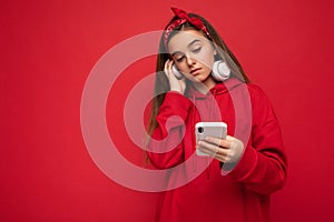 Photo of beautiful concentrated brunette girl wearing red hoodie isolated on red background holding and using smartphone