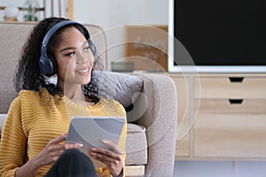 Photo of beautiful dark skin wavy lady homey mood holding digital tablet earflaps listening new audio sitting floor near couch