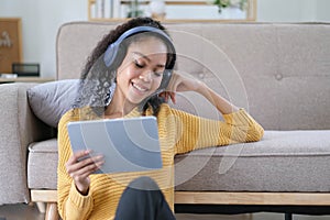 Photo of beautiful dark skin wavy lady homey mood holding digital tablet earflaps listening new audio sitting floor near couch